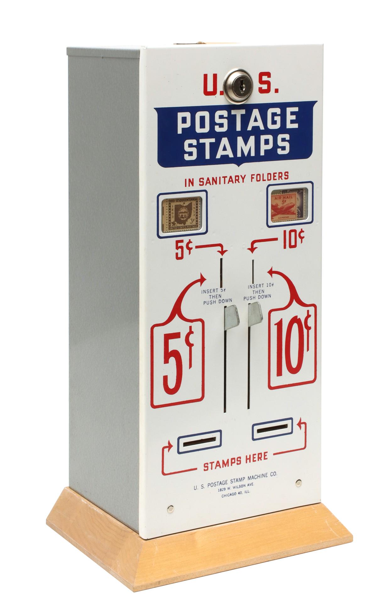 A US POSTAGE STAMPS VENDING MACHINE WITH ORIGINAL BOX: The coin operated vending machine with red, white and blue painted metal case on maple base, the key taped in place, all original and cosmetically mint. The original shipping box retains the original