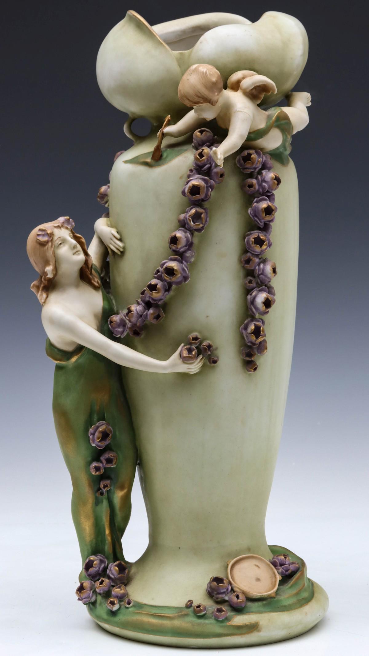 AN IMPERIAL AMPHORA AUSTRIAN ART NOUVEAU VASE GROUPING: The organic vase form of petals above the pinched waist atop baluster form with airborne putti and purple blossom garland flanked by a standing maiden in Classical robe, her tambourine and stray bloss