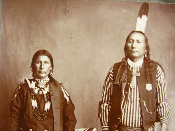 CABINET PHOTO OF TWO PONCA INDIANS C. 1890: Superb albumen image of what appears to be a Ponca husband and wife in tradition dress, except for the husband's giant Christian cross around his neck, law enforcement badge on his vest, watch chain a