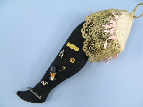 A LADY'S LEG PIN CUSHION WITH ANTIQUE PINS: Measures approximately 12.5 inches, black woolen stocking with lace & silk bows to top, with hanging loop and seven antique pins, including Charlie McCarthy, 'Baby' brooch, a decorative brooch, an 8 b