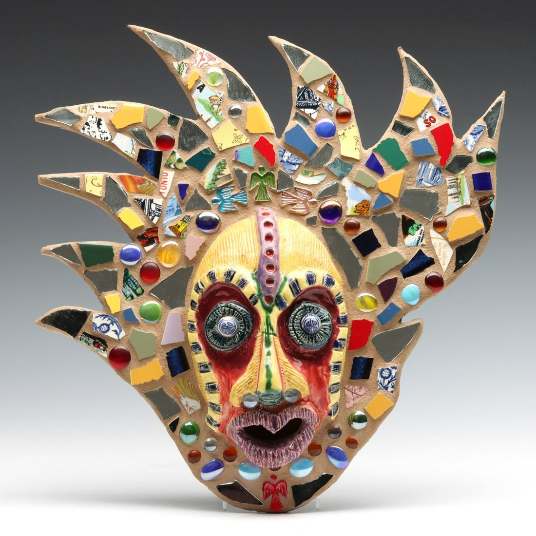 A LATE 20TH CENTURY ARTIST SIGNED MOSAIC MASK: The carved and glazed studio pottery visage set in terra cotta surrounded by pottery shards and iridescent cabochons on scroll-cut Masonite signed by the artist. Measures 15.5 x 15.5 inches. We happil