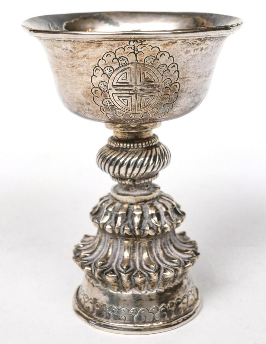 A 19TH CENTURY TIBETAN BUDDHIST YAK BUTTER LAMP: The hand hammered and engraved bowl-form vessel with auspicious symbols on stem with swirled pattern knop above a sculpted band of narrow out-swept petals over flared foot with engraved clouds. No mar