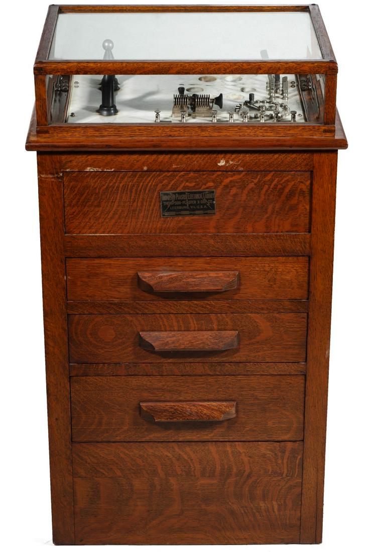 A THOMPSON PLASTER OAK ELECTRICAL CABINET QUACK DEVICE: The circa 1917 violet ray electrotherapy device with marble top under glazed glass top, oak cabinet with drawers, including a divided drawer with blown glass implements and related ephemera and instru