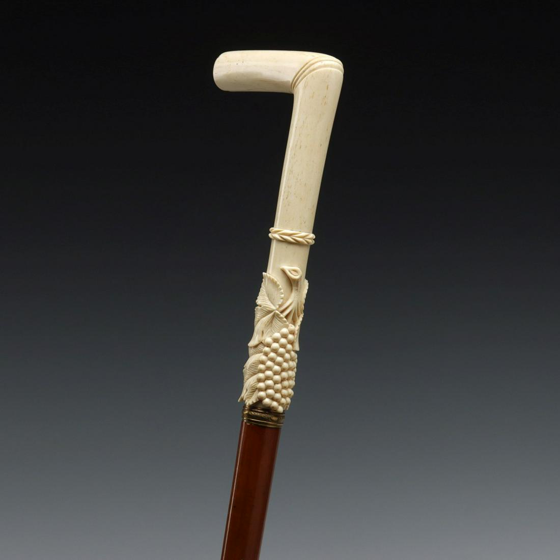 A GOOD 19TH C. 'L' HANDLE CANE WITH CARVED GRAPES: The hollow porous bone handle is crisply carved with grapes in a high relief cluster against detailed leaves above a brass collar with stamped design of rococo swirls on malacca shaft ending in a horn