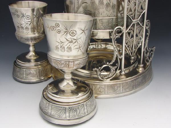 ORNATE VICTORIAN SILVERPLATE WATER TIPPLER: Includes original matching goblets and spill tray, bases marked "Pairpoint, Quadruple Plate." Stands 20.5 inches, 9 x 12 inch base, pitcher stands 12.5 inches.