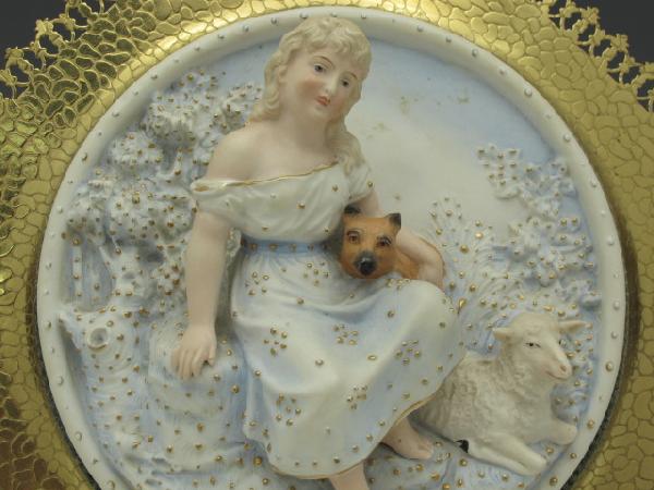 AN UNUSUAL VICTORIAN BISQUE PLAQUE: Hand tinted 3D plaque with girl sitting with fox and sheep, gold stipples throughout, mounted to polished brass free-standing frame with curled corners and cut-out edges, measures 11 inches across.