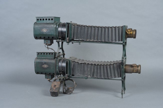 A BRENKERT SLIDE PROJECTOR MODEL D1: Stackable slide projectors, two here, each with Brenkert Light Projection Co spec tags affixed to sides. Full bellows, and power cord with switch box. Focusing knobs on both lenses. Measures 20.5 x 30