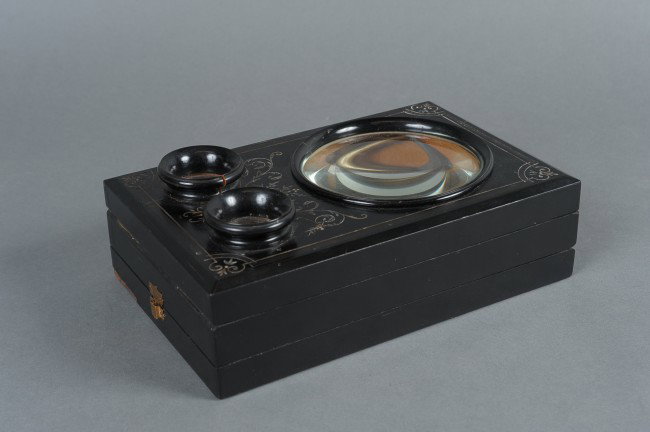 A GERMAN GRAPHOSCOPE BY C. ECKENRATH, BERLIN: 19th century graphoscope with carved and black painted wood face, large magnifier lens, two stereoscopic lenses bleow. Folding table base with C. Eckenrath, Berlin spec plate to leg. Folding picture h