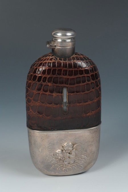 AN UNGER BROS 925 SILVER CHERUB FLASK: Glass flask with hinged lid, alligator skin pattern leather cover, and Unger Brothers marked 'Sterling 925 Fine' marked sleeve with embossed cherub and woman in the clouds design. Measures 6.25 x 3 in