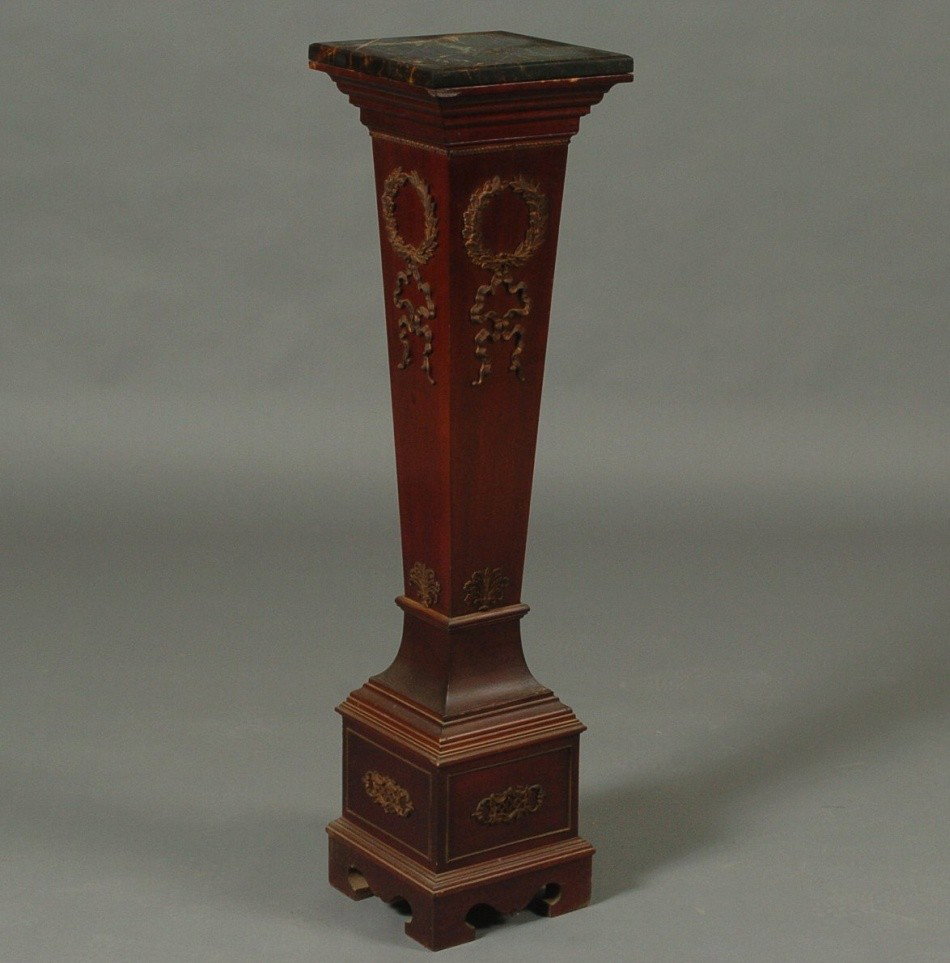 MAHOGANY NEOCLASSICAL-REVIVAL STYLE PEDESTAL: Square black marble top over stepped projected molded cornice over tapering square form, frieze with gilt brass motif of ribbon and bound laurel swag over scrolling foliate, with applied gilt brass de