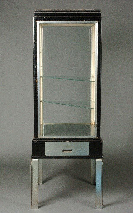AN ART DECO DISPLAY CASE MANNER OF BEL GEDDES: A machine age skyscraper design reminiscent of the work of Norman Bel Geddes. Maker's label of Charles Arnao Company inside one drawer. Two drawers and two slide out shelves on each side. Iron legs an