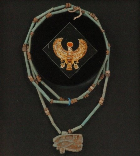 TWO EYE OF HORUS AMULETS, ONE WITH NECKLACE: A soapstone carved Eye of Horus amulet, greenish-gray, matted and framed, 1 x 1.25 amulet in a 17.25 x 10.25 frame; and a turquoise-colored faience amulet on a pipe and disc bead necklace, 1.25 x .75
