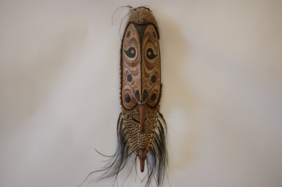 IATMUL SEPIK RIVER MASK & A WOOD CARVING, NEW GUINEA: A ceremonial mask, also called a homicide mask, of wood, clay, shells and feathers with a painted face and cowry shell eyes. Beard and edges are decorated with shells embedded in clay. A carved beak e