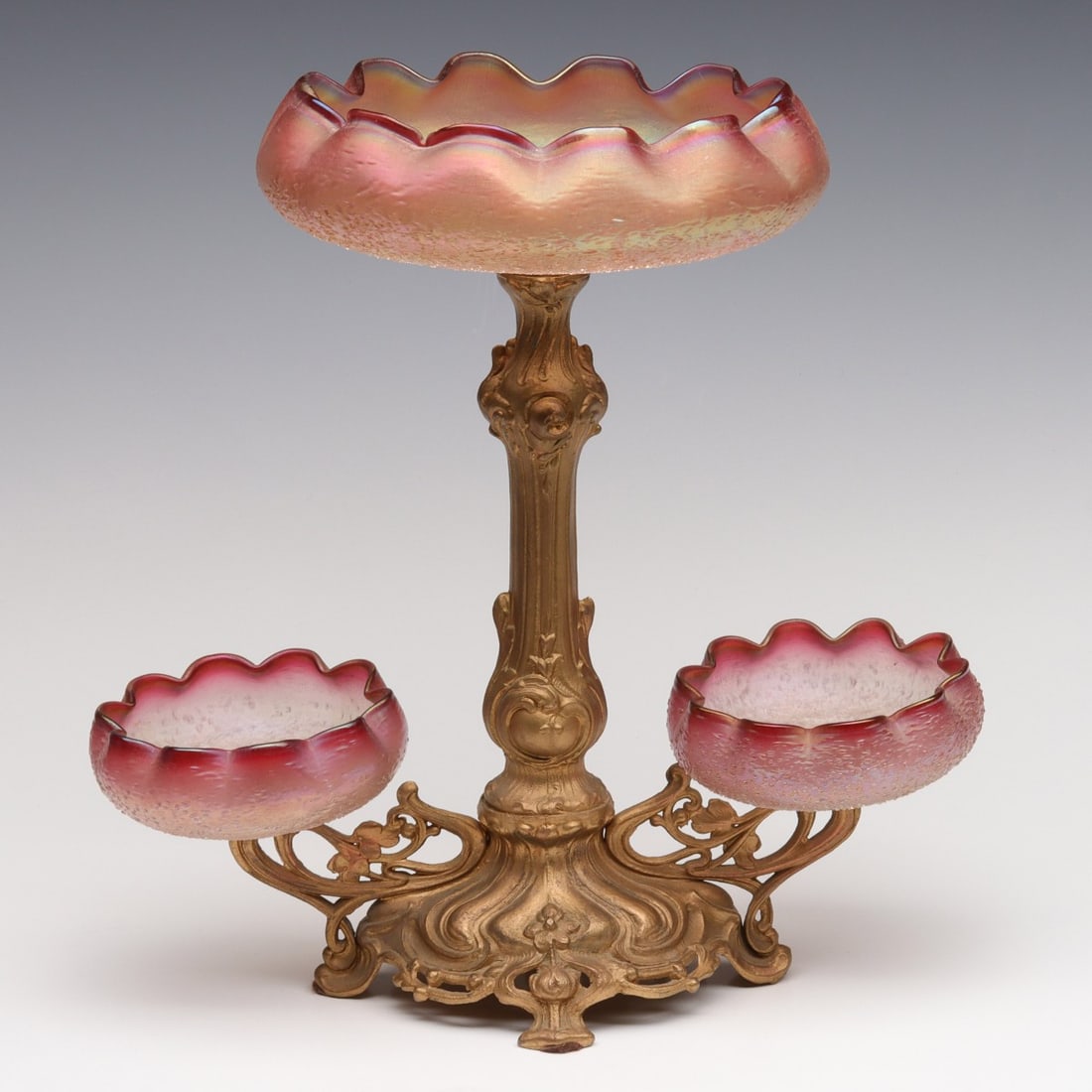 A JUGENDSTIL INFLUENCE EPERGNE WITH AUSTRIAN ART GLASS: The white metal stand with sinuous lines is set with three matching fritted rubina glass bowls with crimped edges and iridescent finish. No signatures found. It is unclear whether or not the glass pie