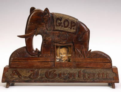 A 1930s FOLK ART CARVING ATTRIBUTED TO A KANSAS INMATE: The solid walnut carving of a G.O.P. elephant on long trapezoidal platform is centered by a picture frame and lettered along the base in bas relief. A pop-up elephant figure backs the picture frame.