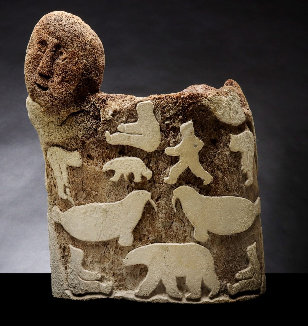 A MASSIVE EARLY 20TH CENTURY INUIT WHALEBONE CARVING: The large half-round section of bone displays Native subject matter in three different techniques. In addition to the two-sided figure that surmounts the piece, seals and ptarmigans are incised on one