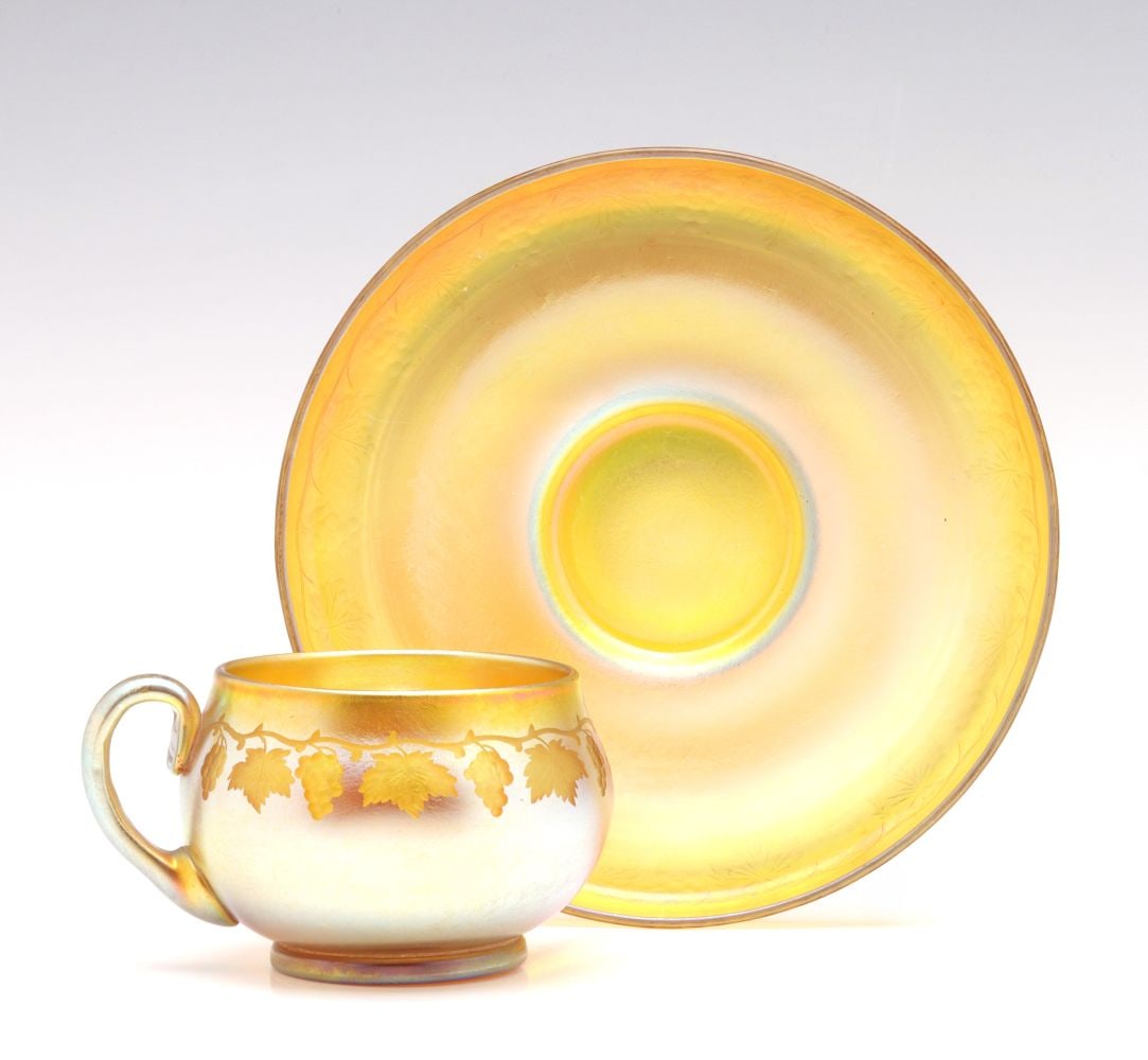 A TIFFANY FAVRILE CABINET CUP SAUCER SET WITH ENGRAVING: The small gold favrile cup and saucer displays a very fine iridescent finish and intricately, executed intaglio engraving of an encircling grapevine with hanging clusters and leaves. The base with whe