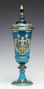 A MOSER BLUE ART GLASS DECORATED POKAL WITH COVER: The large covered vessel is decorated with a heraldic-like motif of child with doves rising from flowering acanthus swirls. Measures 17 x 5.25 inches. Without proof of exemption, be aware that