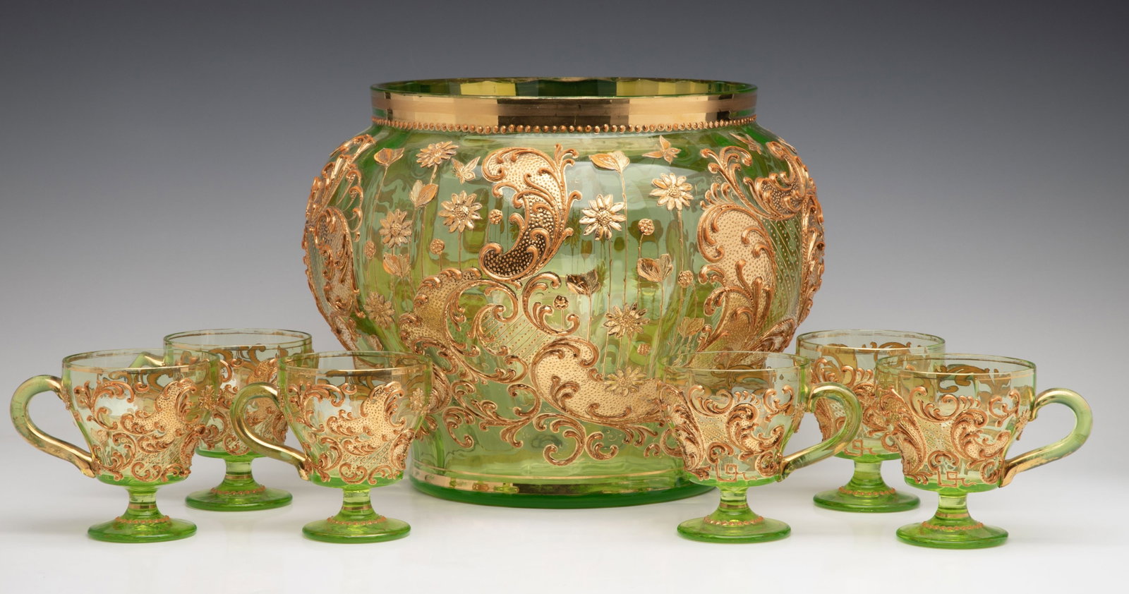 A BOHEMIAN GLASS PUNCH BOWL AND CUPS WITH THICK ENAMELS: The green glass is embellished in thick gold rocaille and rococo swirls interspersed with flower heads on this stems. Bowl measures 9 x 11.5, cups are 3.625 x 3.25 inches each. Without proof of exempt