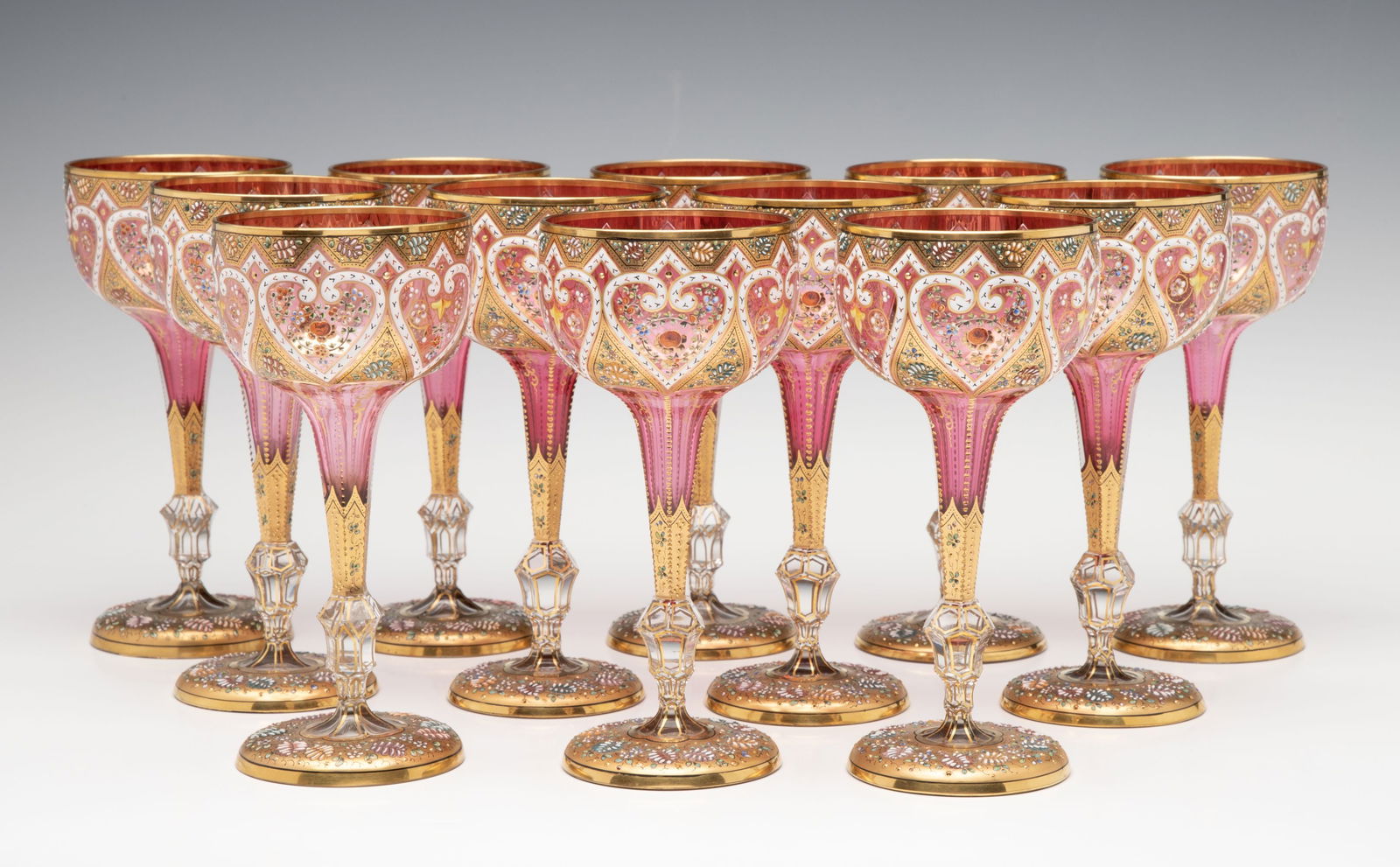TWELVE ELEGANT GILDED AND ENAMELED MOSER WINE GOBLETS: Each cranberry glass goblet is lavishly decorated in Persianate enamels and rich gilding at the rim and on the four sided stems with tiny beading and zipper cut facets. Each colorless glass foot is cu