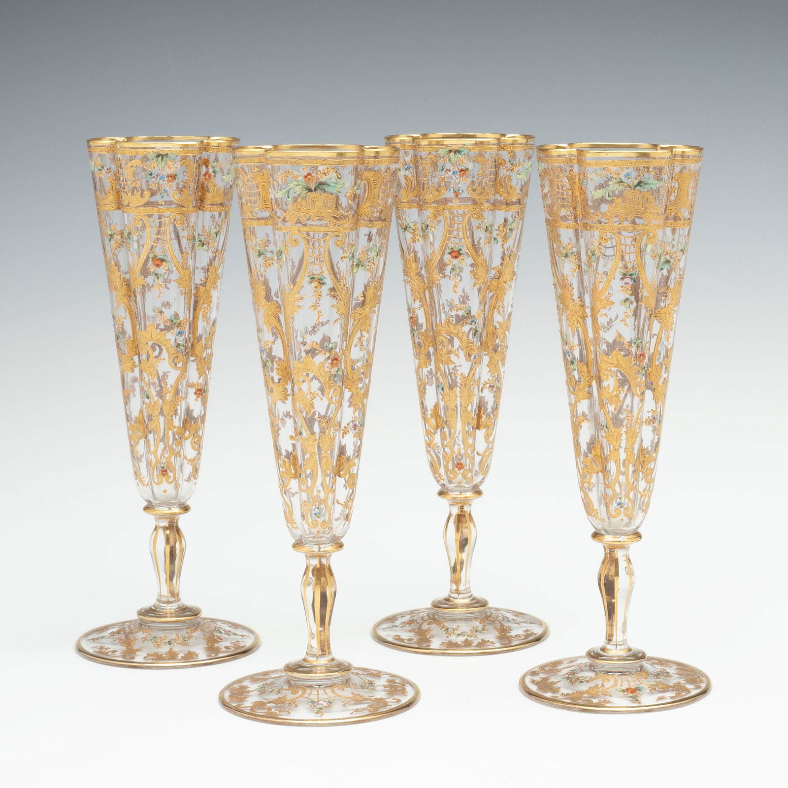 FINE AND THINLY BLOWN MOSER CRYSTAL FLUTES WITH ENAMEL: Ludwig Moser Late 19th Century Each thinly blown quatrefoil shape glass is decorated in relief enamels of gold rocaille and rococo swirls amongst petite colorful enamel flowers and raised on a short s