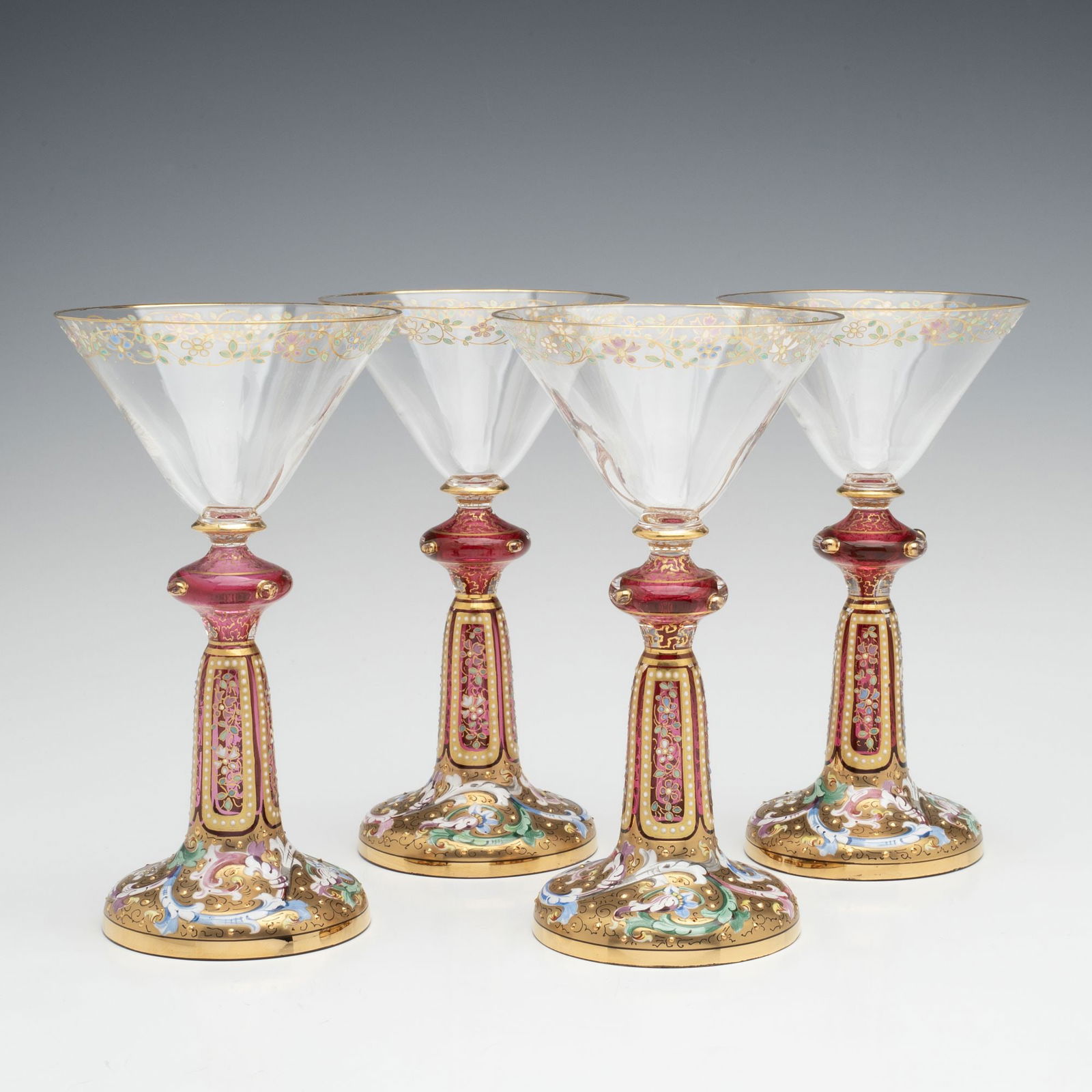 FOUR VERY UNUSUAL GILT AND ENAMEL MOSER WINE GOBLETS: Ludwig Moser Late 19th Century Each stem is comprised of an inverted conical crystal bowl subtly decorated with a simple band of enamel floral meander, and raised on a cranberry stem with flattened kn