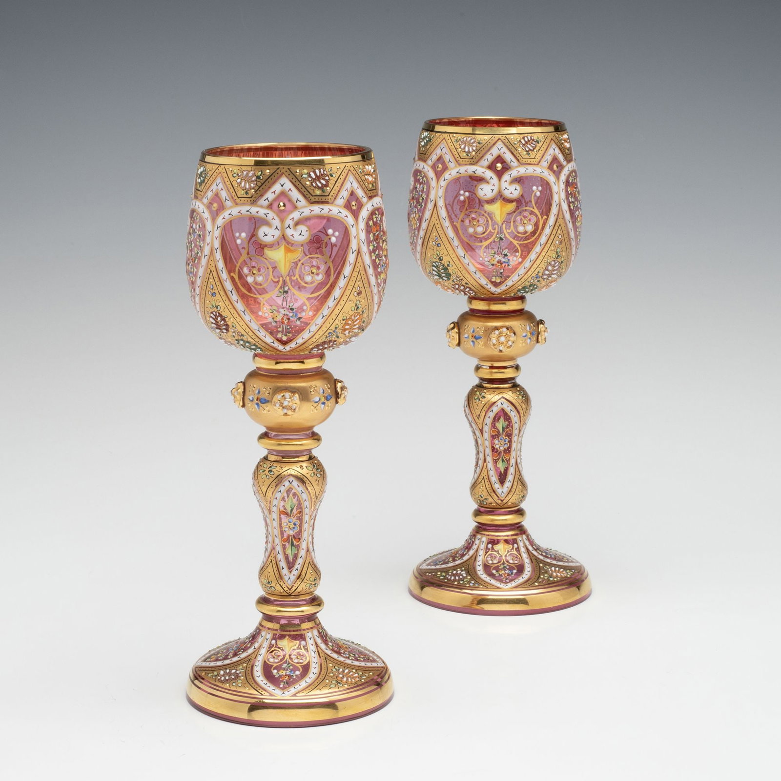 MOSER ISLAMIC INFLUENCE GILT AND ENAMELED WINE GOBLETS: Ludwig Moser Late 19th Century Each goblet is lavishly decorated in rich gold and colorful enamels incorporating motifs of Islamic tradition, and four beaded prunt applications, all on light cranberry