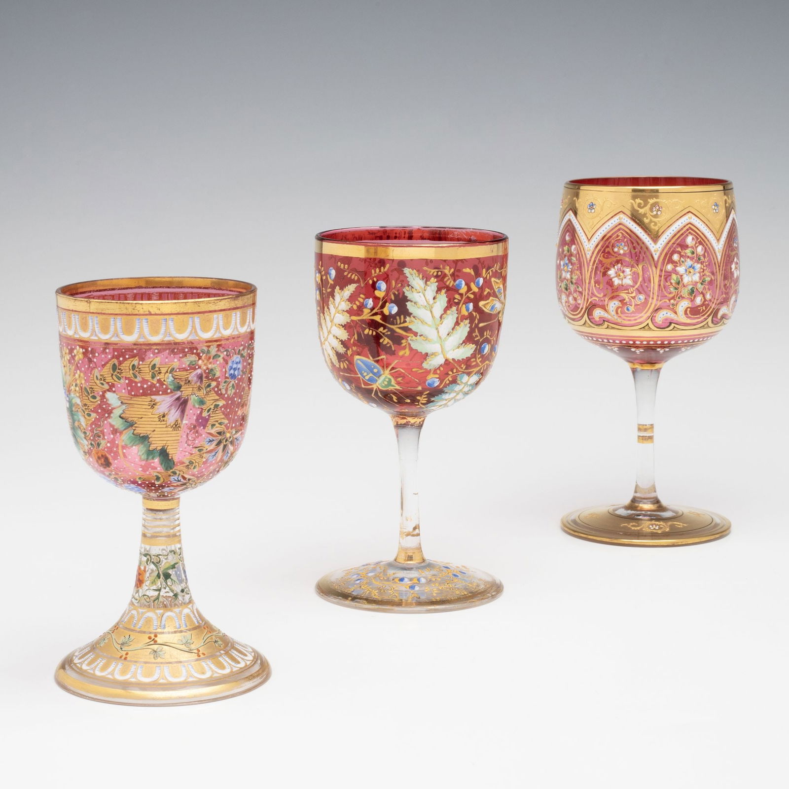 THREE UNIQUE MOSER CRANBERRY GLASS SMALLER WINE STEMS: Ludwig Moser Early 20th Century Each having a beautifully decorated cranberry glass bowl embellished in multi-colored enamel designs incorporating foliates, florals, berries, insects and traditional I
