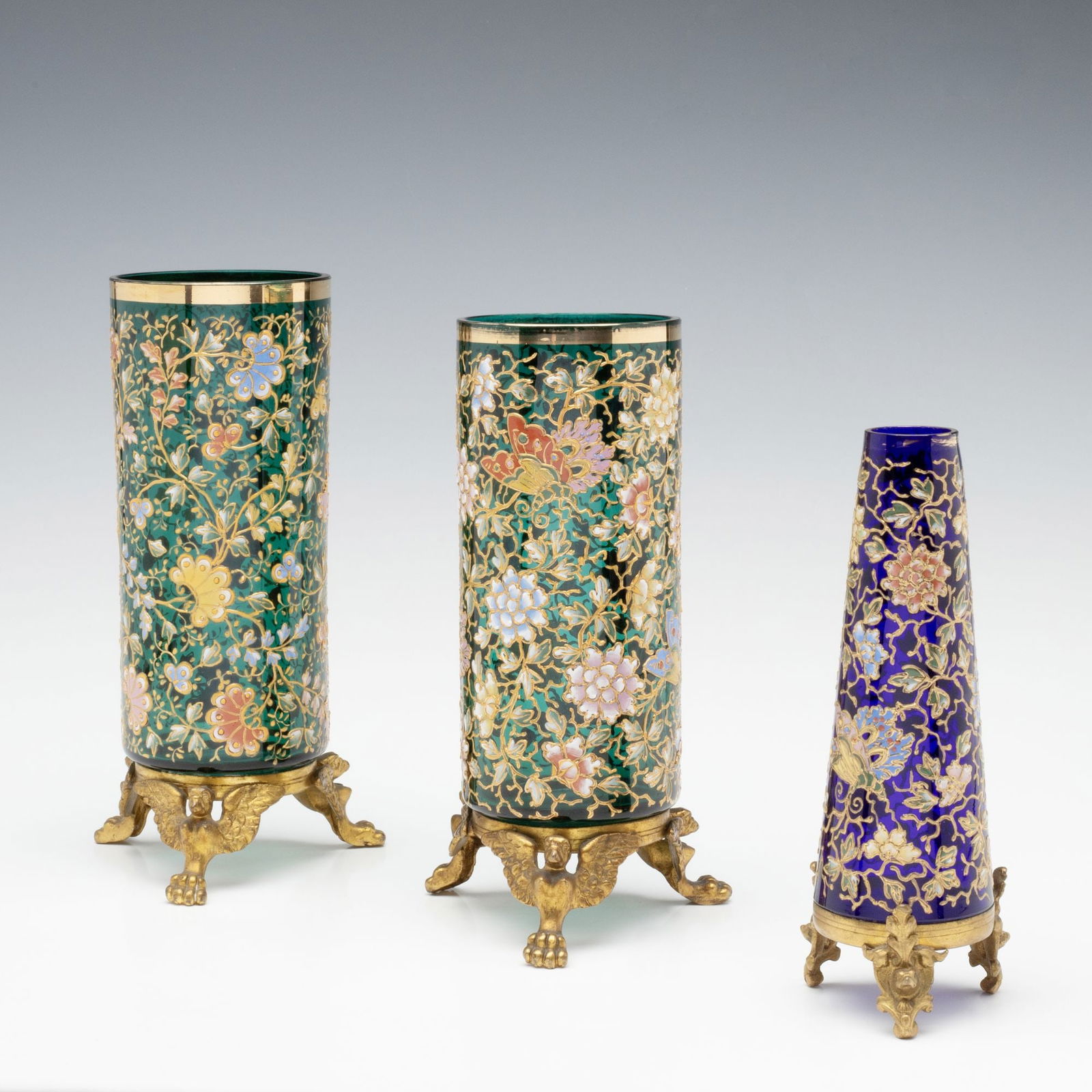 ENAMEL & GILT DECORATED MOSER ART GLASS IN METAL MOUNTS: Ludwig Moser Early 20th Century Each of the three unusual examples is raised on a gilded base and covered in elaborate, colorful hand painted enamel decoration of swirling vinery, flower heads, and bu