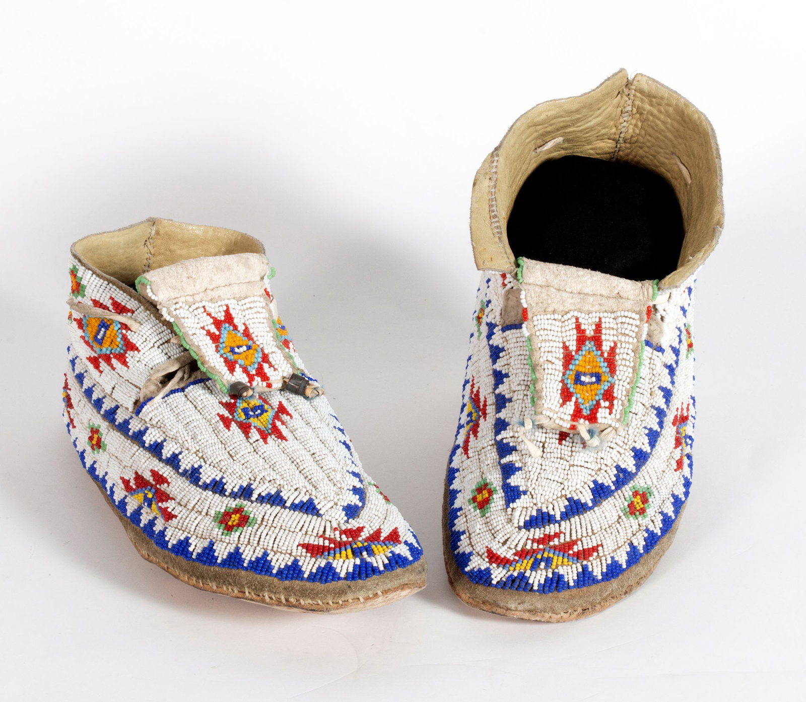 A PAIR RED, WHITE, BLUE PLAINS INDIAN BEADED MOCCASINS: The circa 1940 pair are decorated in medium blue, red, yellow, green, white and turquoise glass beads stitched in complex geometric designs reminiscent of those found in Navajo weavings, all above the