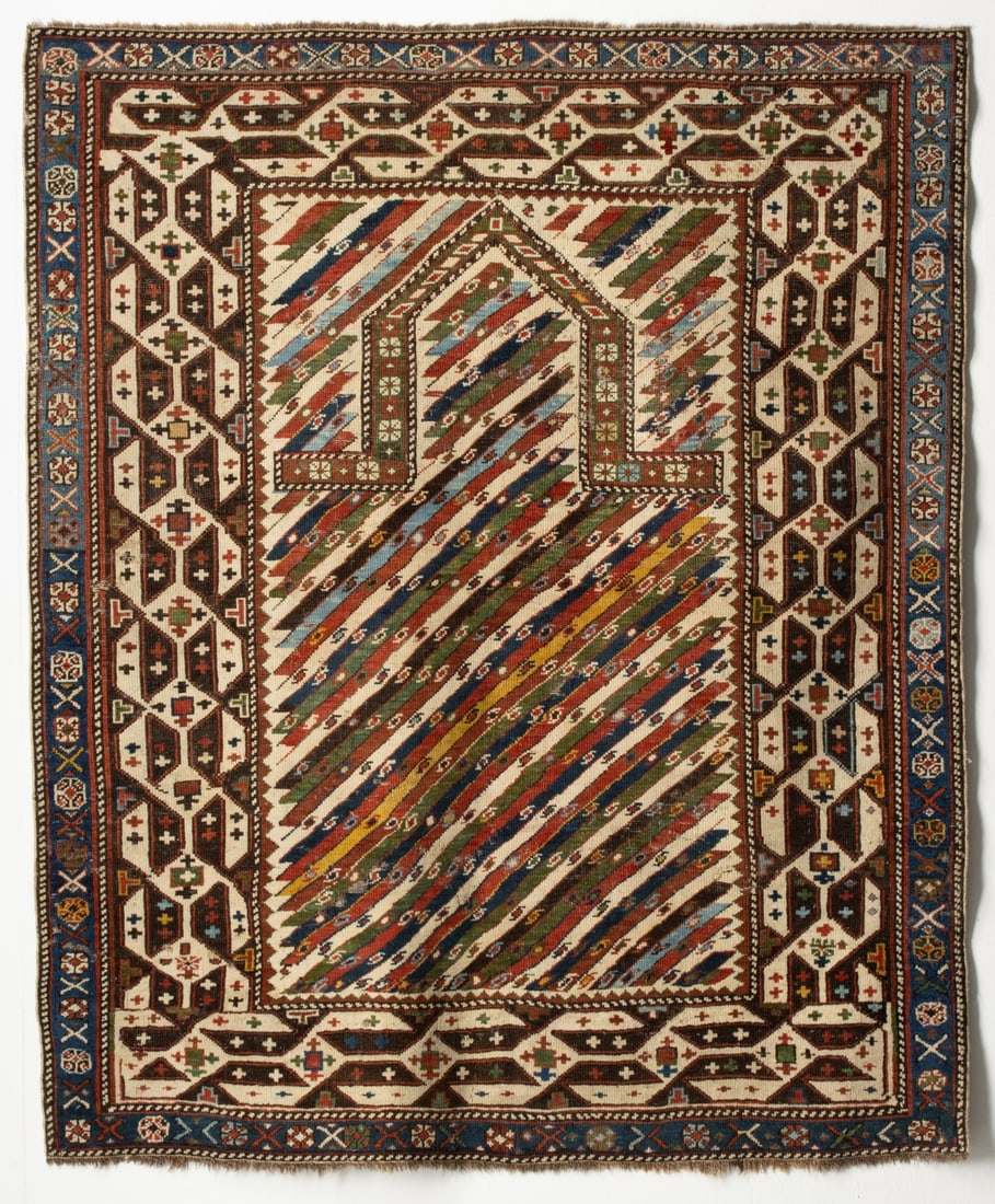 UNUSUAL SHIRVAN PRAYER RUG, CIRCA 1880: The ivory mihrab with diagonal polychrome striped canes, below a pointed arch, similar striped spandrels, within a dark blue border enclosing a continuous running dart border, a pale blue flower head