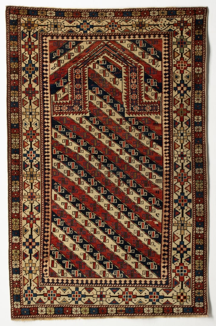 A SHIRVAN PRAYER RUG, EAST CAUCASUS, LATE 19TH CENTURY: With alternating red ivory and blue diagonal stripes enclosing rectangular hooked flower heads, below a pointed arch, similar diagonal banded spandrels, within an ivory stepped medallion and bracket v