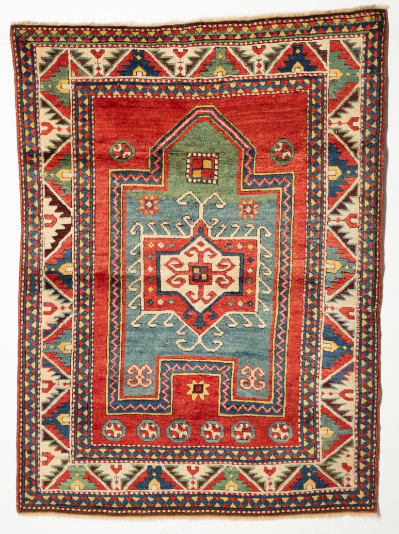 A FACHRALO KAZAK PRAYER RUG, CIRCA 1880: The shaded red field with an abrashed green and pale blue mihrab enclosing a red and ivory hooked rectangular medallion, supported by wheel flower heads, within a polychrome reciprocal diagonal serrat