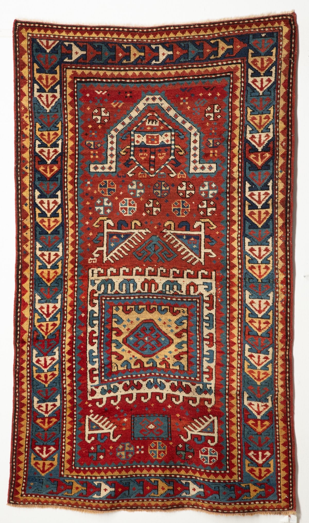 A FACHRALO KAZAK PRAYER RUG, CIRCA 1880: The brick red field with a hooked ivory square medallion, hooked blue spandrels, supported by assorted flower heads below a flattened pointed arch, red flower head spandrels, a blue border enclosing h