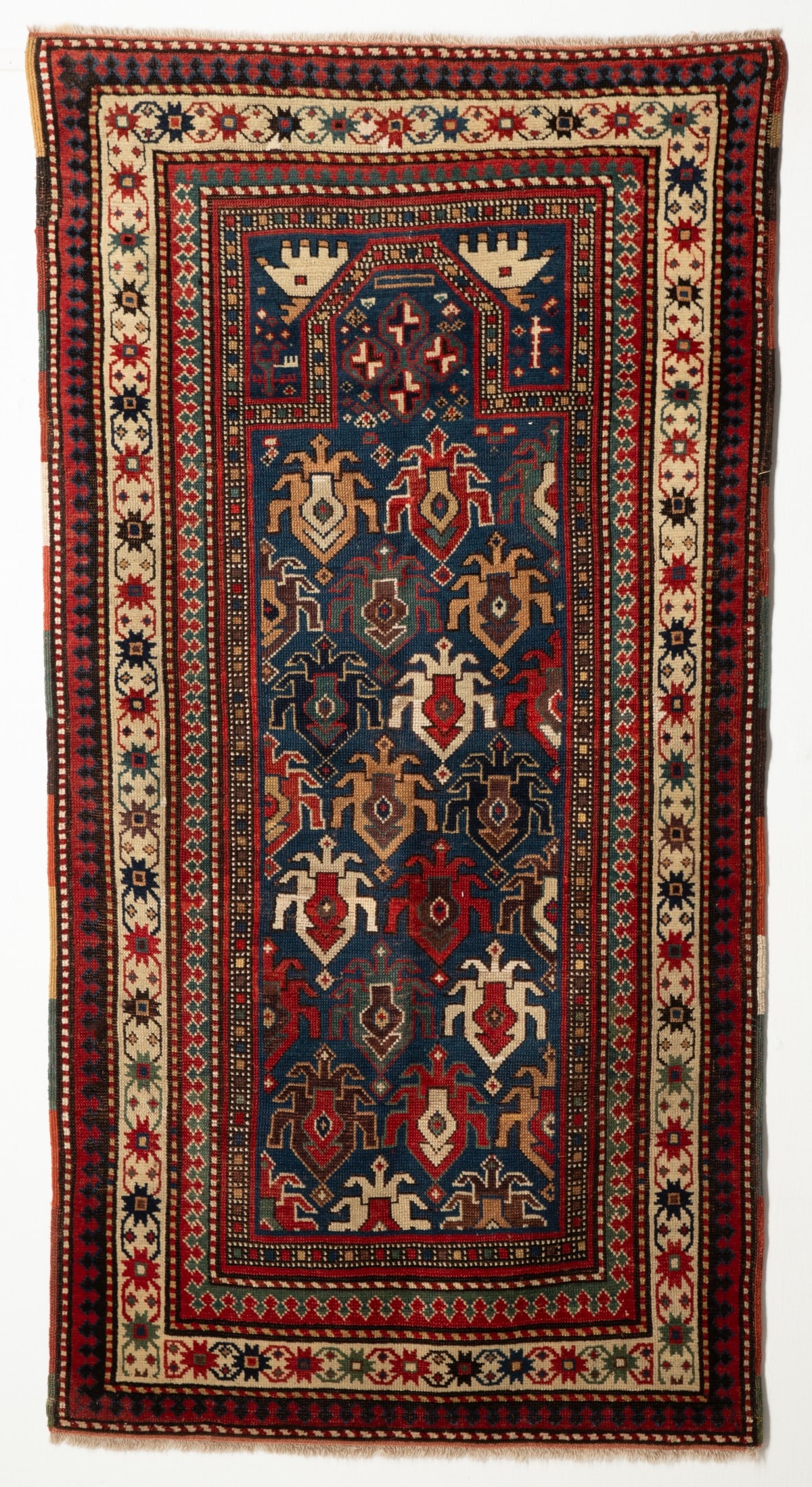 AN UNUSUAL KAZAK PRAYER RUG, CIRCA 1880: The blue field with allover offset hooked shield palmettes below a flattened pointed arch, blue hand spandrels within an ivory star flower head continuous bracket vine border, reciprocal medacyl twin