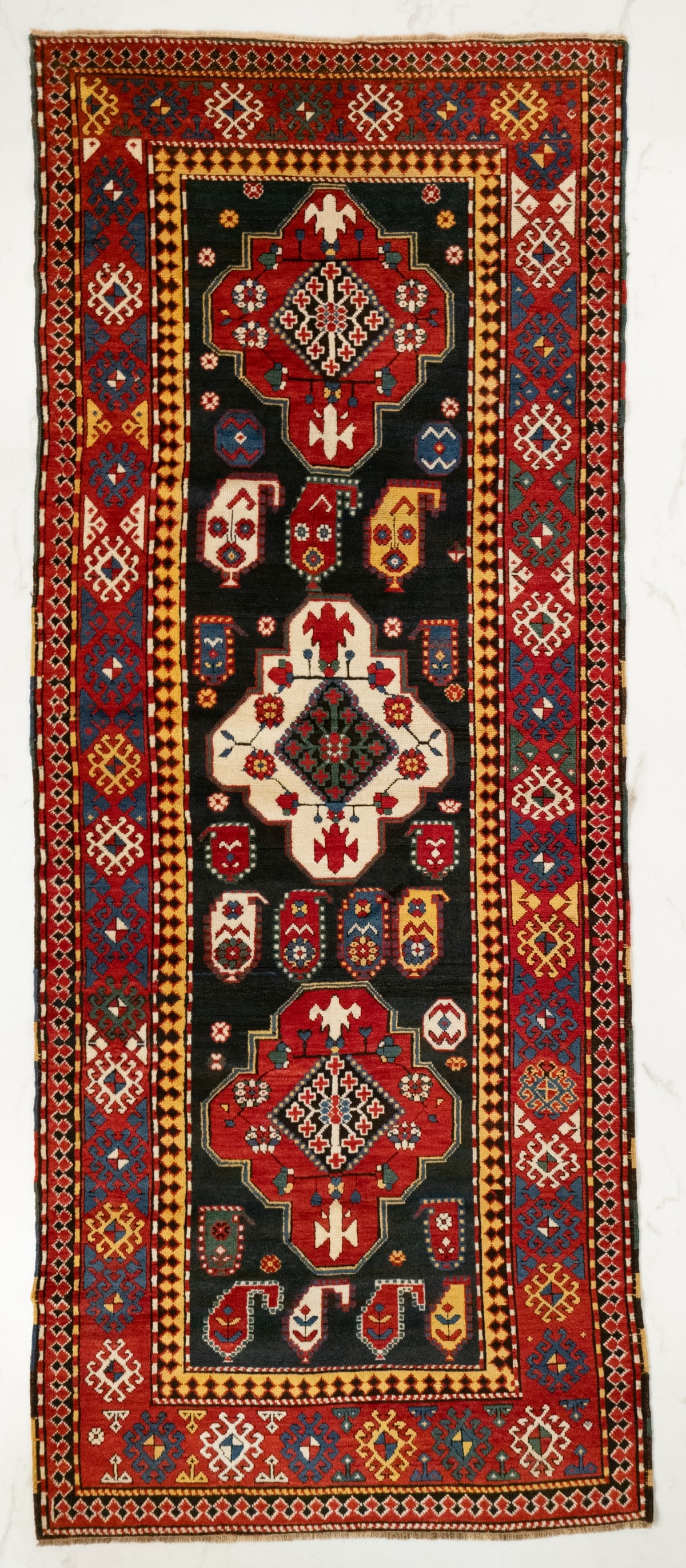 A GENDJE KAZAK LONG RUG, EARLY 20TH CENTURY: The dark blue field with a central ivory stepped medallion accompanied by two similar red medallions supported by rows of boteh and flower heads, within a red border enclosing continuous hooked rhombu