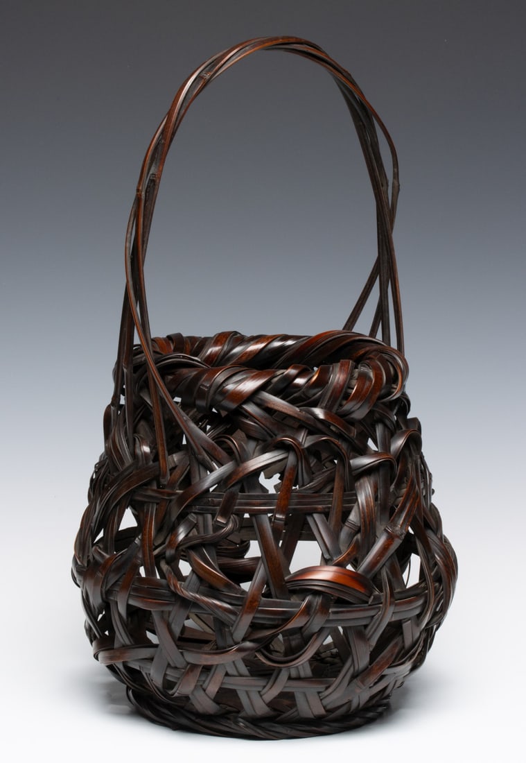 TANABE CHIKUUNSAI I BAMBOO IKEBANA BASKET, ADAIR ESTATE: The globular form is composed of broad, heavy and random bamboo strips that overlap and interlock in a dramatic way. The wide, rolled rim is constructed with thick, bundled bamboo and the handle is fo