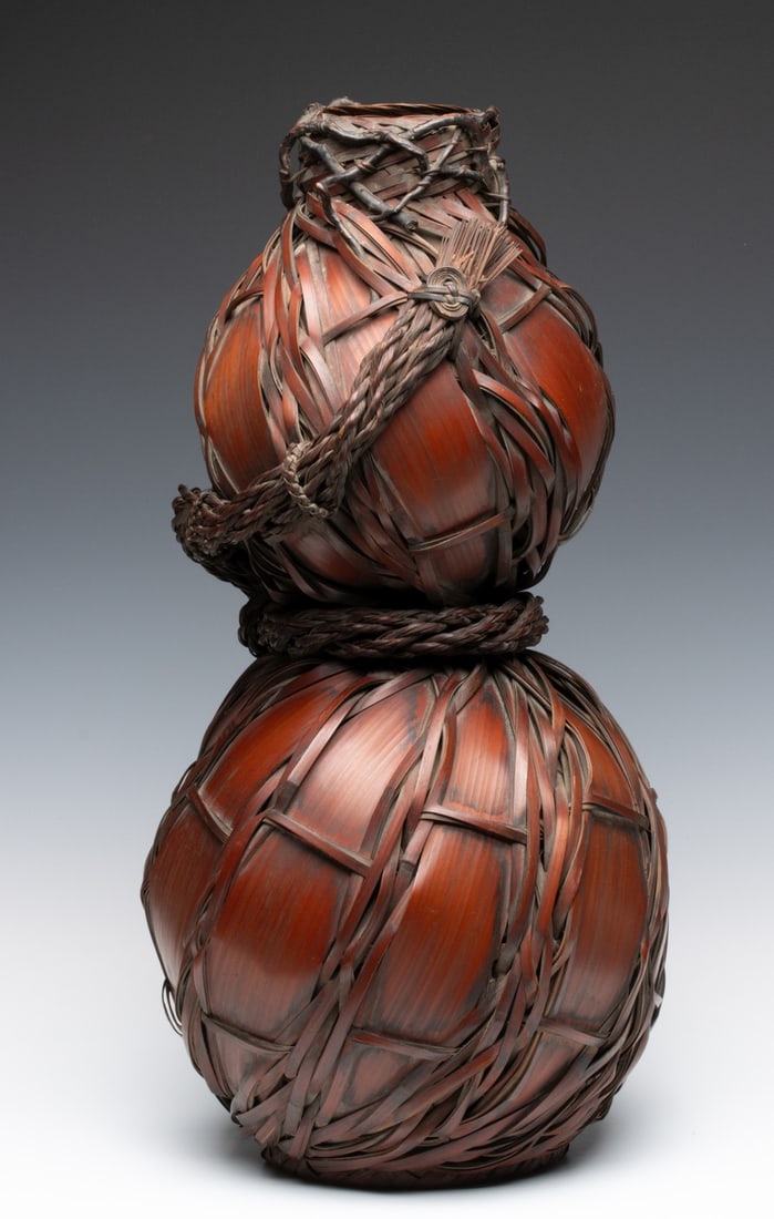 A JAPANESE DOUBLE GOURD IKEBANA BASKET, ADAIR ESTATE: The sculptural double-gourd form composed from broad, overlapping randomly woven bamboo splints is wrapped and cinched at the waist with a neck finished by a rustic wrap of gnarled bamboo stalks aroun