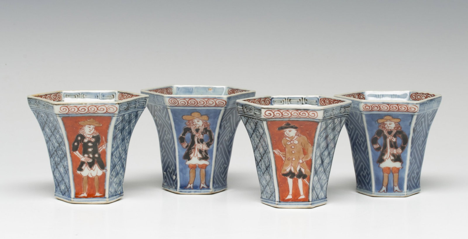FOUR MEIJI JAPANESE KUTANI CUPS WITH DUTCH TRADERS: A set of four Japanese Kutani porcelain six-sided cups, Meiji period, each decorated in iron-red, blue, and gilt with standing Dutch trader figures reserved within shaped panels, the sides with geomet