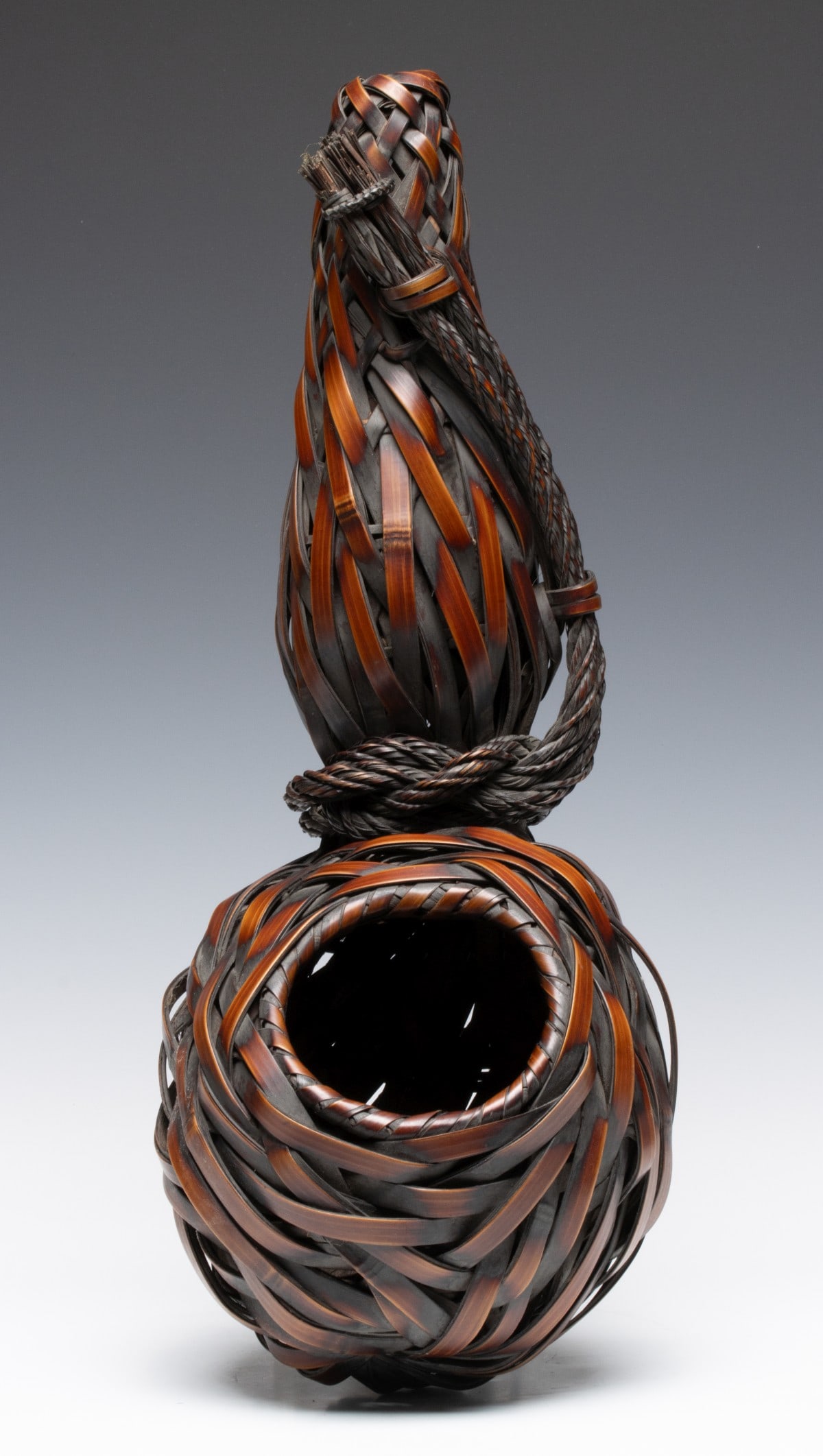 A HANGING JAPANESE BAMBOO IKEBANA BASKET, ADAIR ESTATE: The hanging double gourd basket is constructed with bamboo splints that swirl around the body cinched at the waist by a thick, hand-braided bamboo rope to create the double gourd form while extending