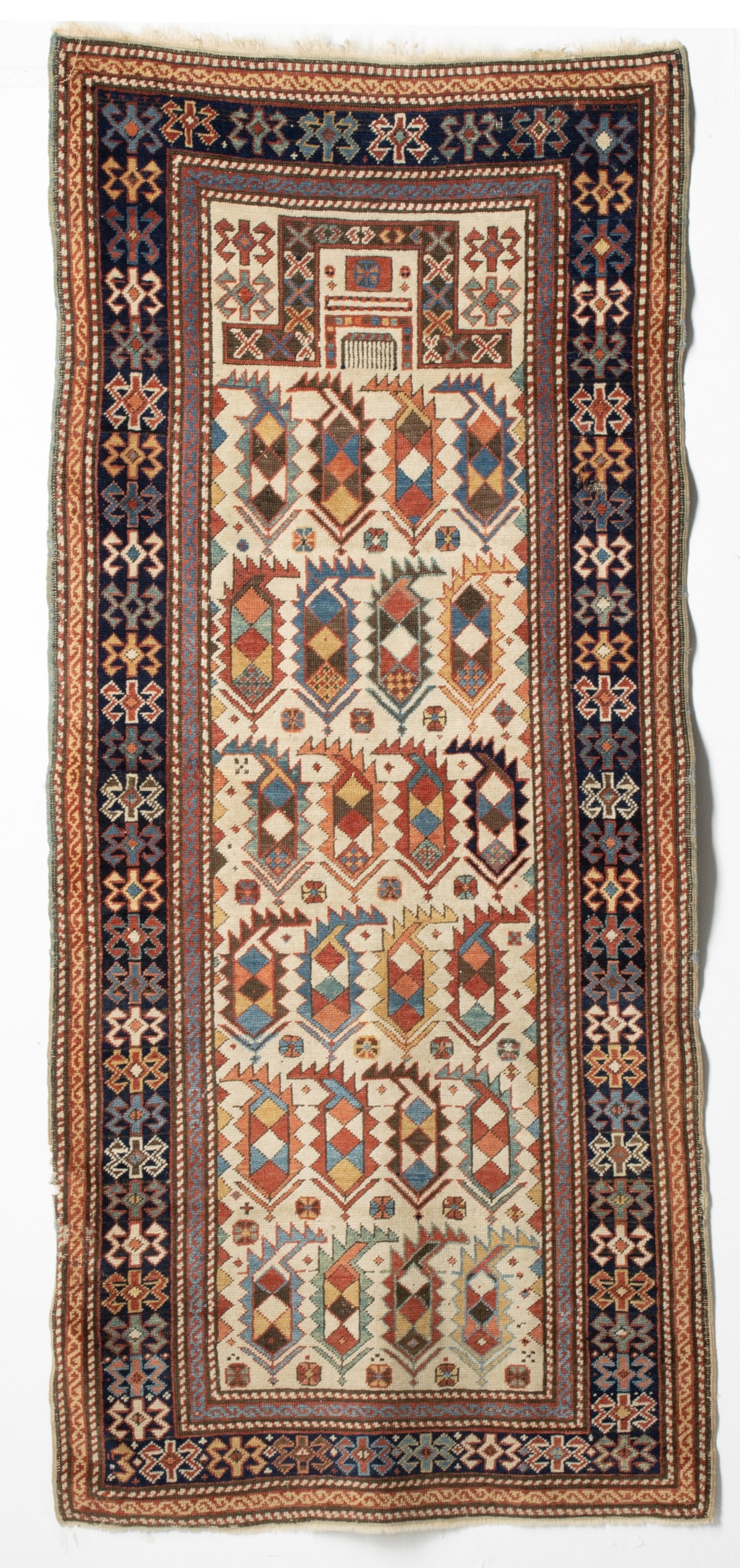 SHIRVAN PRAYER RUG, EAST CAUCASUS, CIRCA 1875: The ivory mihrab with rows of 'in and out' polychrome serrated harlequin boteh below a flattened arch enclosing a comb dais, hooked medallion ivory spandrels, within a dark blue border enclosing