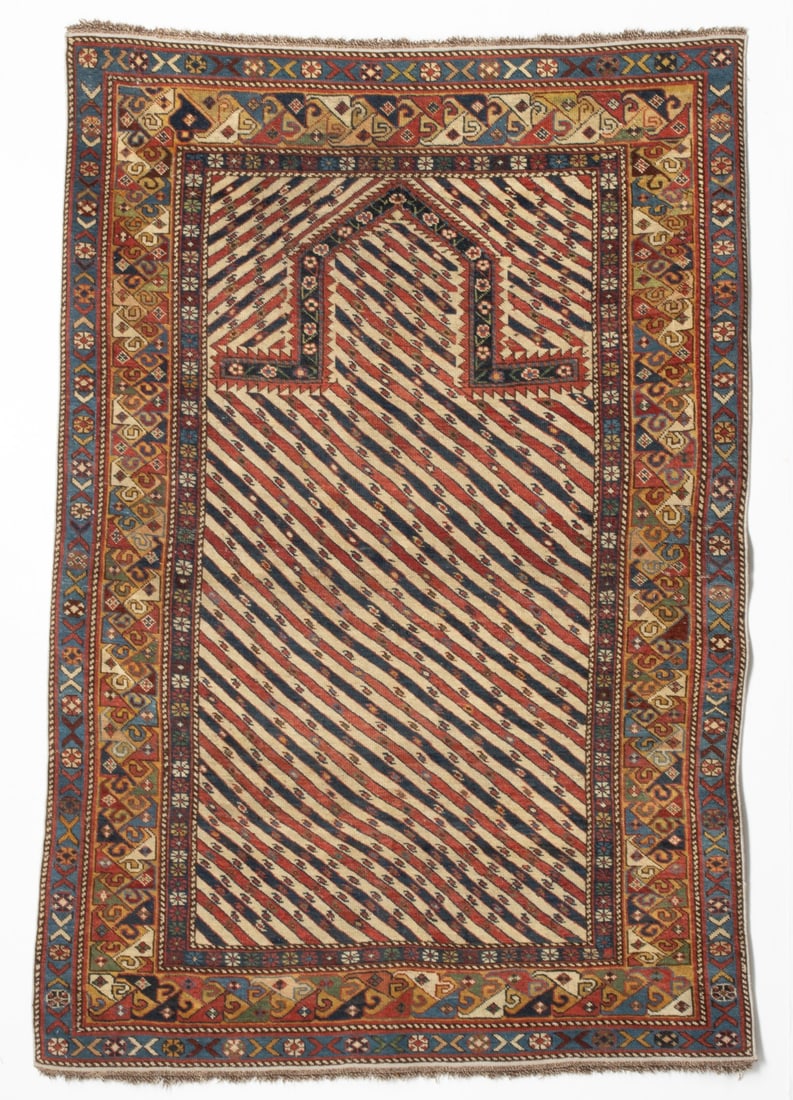 SHIRVAN PRAYER RUG, EAST CAUCASUS, LATE 19TH CENTURY: The ivory mihrab and spandrels with all over alternating red and blue diagonal bands enclosing continuous tiny flower heads supporting a pointed arch within a saffron border enclosing continuous linke