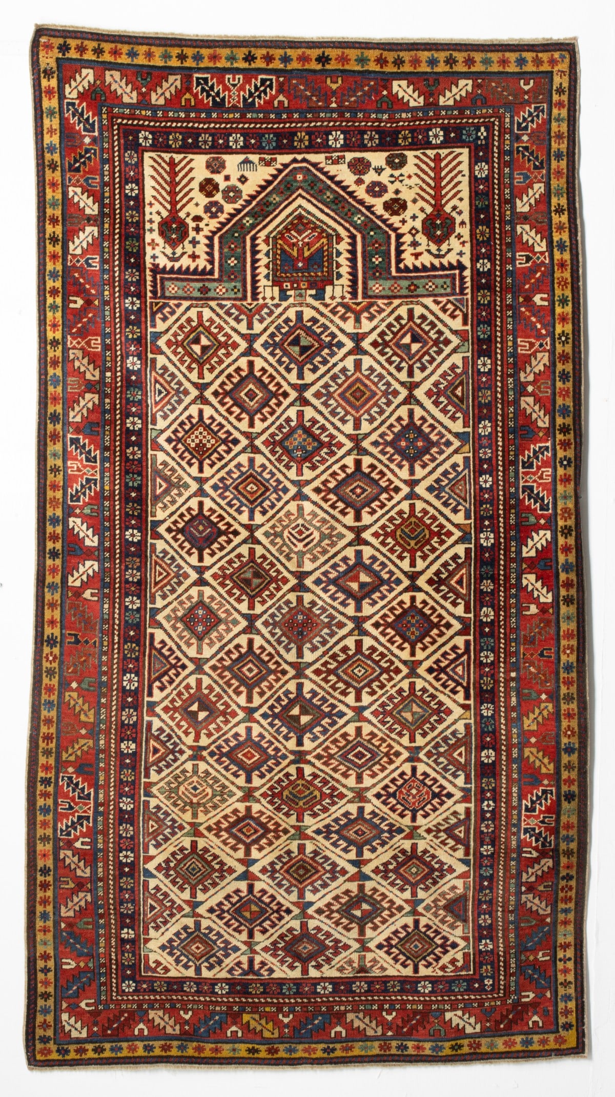 SHIRVAN PRAYER RUG, EAST CAUCASUS, CIRCA 1880: The ivory mihrab with allover offset hooked lozenge medallions enclosed within a rhombus tile lattice, below a pointed arch, ivory spandrels enclosing a pair of mirrored chevron plants and flower head