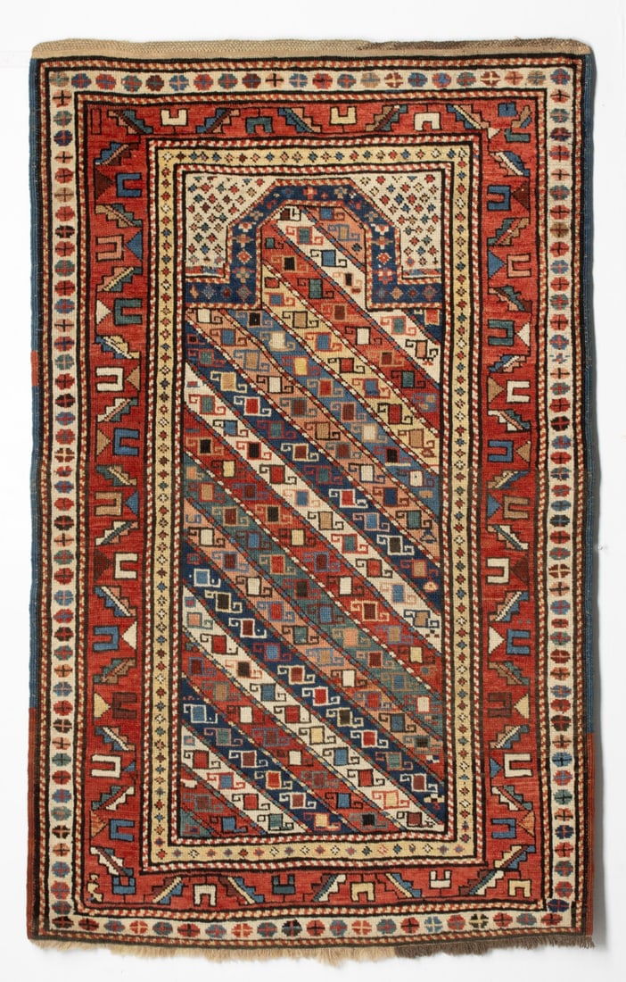 GENDJE PRAYER RUG, CIRCA 1880: The polychrome diagonal banded mihrab enclosing continuous hooked square flower heads, below a flattened chamfered arch, ivory spandrels, within a red serrated leaf and calyx border, an ivory continuo