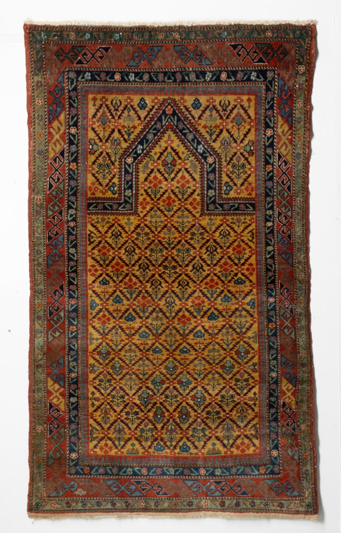 DAGHESTAN PRAYER RUG, LAST QUARTER 19TH CENTURY: The deep saffron mihrab with an allover serrated leaf lattice enclosing multifarious flowering plants below a pointed arch, similar deep saffron spandrels within a red border enclosing continuous diag