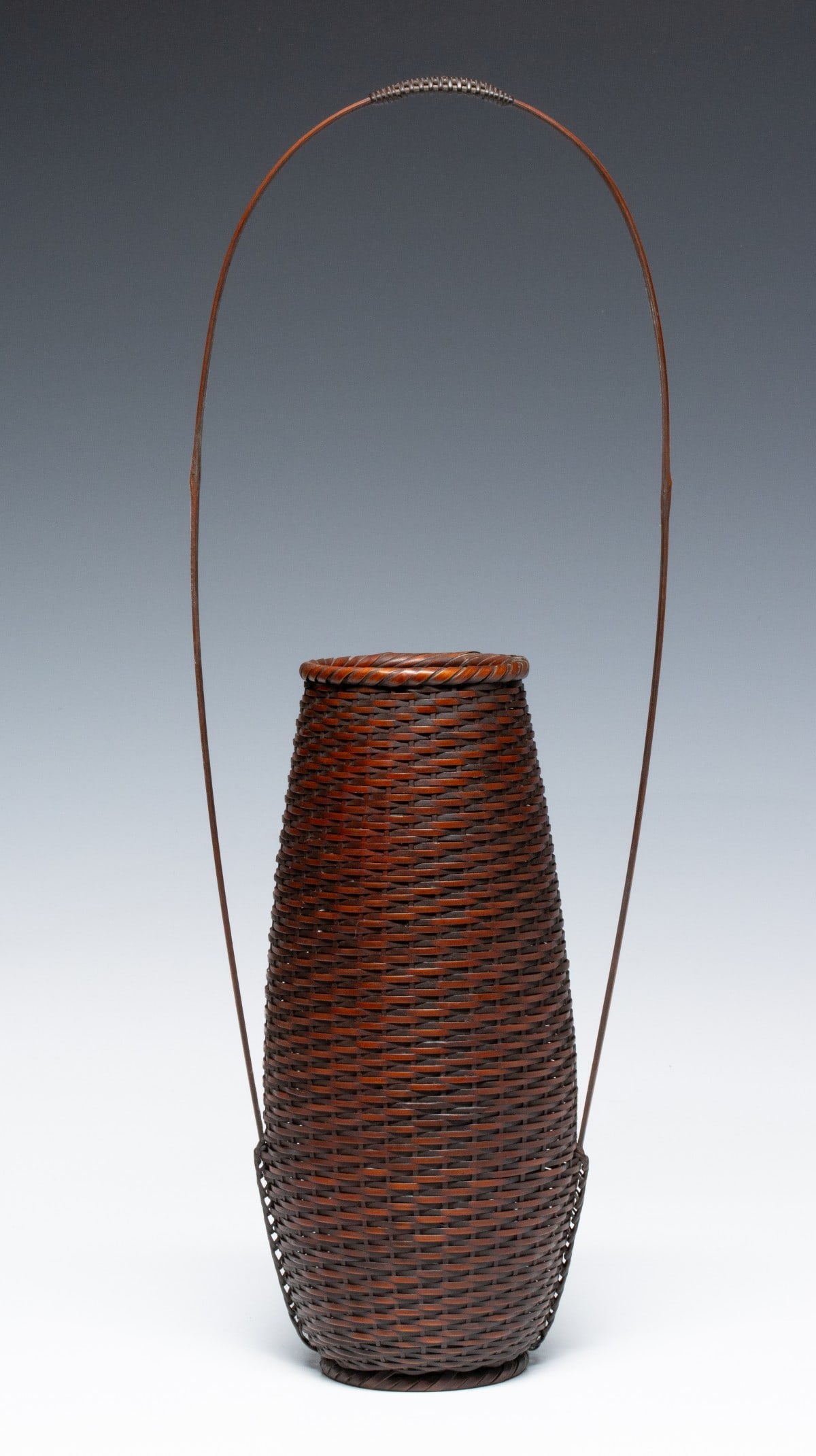A FINE JAPANESE BASKET SIGNED CHIKUYOSAI, ADAIR ESTATE: The elegant, understated basket for ikebana in rich, dark-amber patina is constructed in a meticulous horizontal twill weave and enhanced by an exceptionally tall, airy handle that creates an