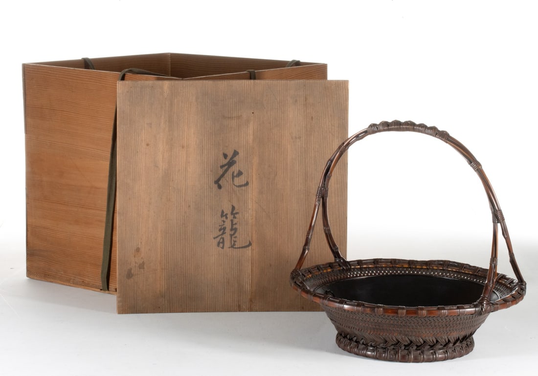 TANABE CHIKUUNSAI I HANDLED BAMBOO BASKET, ADAIR ESTATE: The vessel is constructed in a highly refined twill plaiting that transitions at the base into a striking decorative band of overlapping diagonal slats. The basin has an overhead handle composed of la