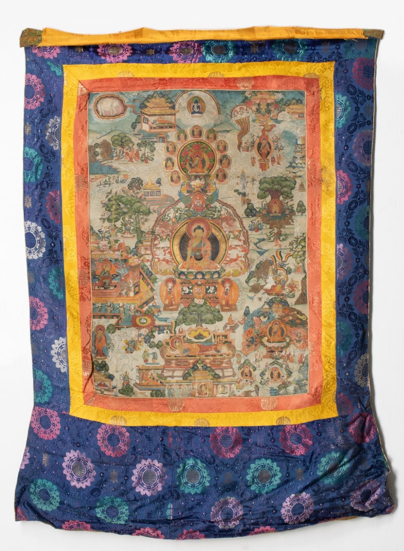 A 19C. TIBETAN THANGKA OF THE LIFE OF SHAKYAMUNI BUDDHA: The sacred scroll painting depicts the life and narrative cycles of Shakyamuni Buddha seated in dhyanasana on a multi-colored lotus throne over a decorative pedestal. His right hand is extended in bhu