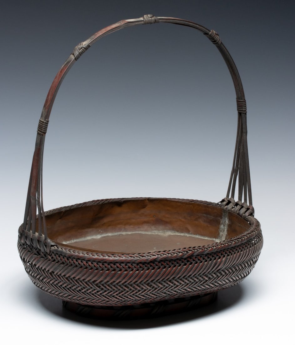 A FINE JAPANESE FLOWER ARRANGING BASKET, ADAIR ESTATE: The shallow rounded form is skillfully woven from fine strands of bamboo in a dense bi-tonal twill finished in a tightly woven rim joining the high, arched handle composed of layered bamboo slats that