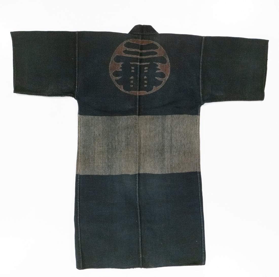 AN INDIGO JAPANESE FIREMAN'S COAT W/CREST, ADAIR ESTATE: With circular brigade emblem to the back and presumably related insignia to the lapels. The bold graphic design and identifying insignia likely relate to a specific firefighting company or regional